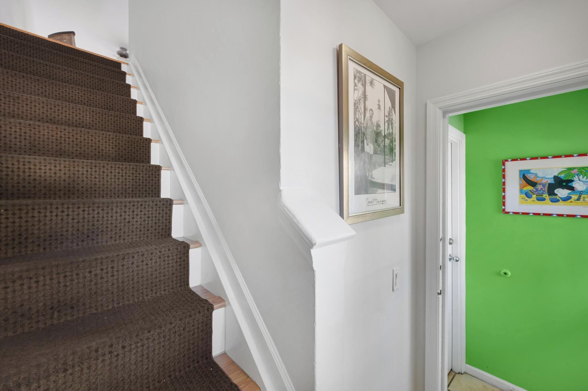A staircase with a brown carpet and white walls, a framed picture on the wall, and a green door with a colorful border.