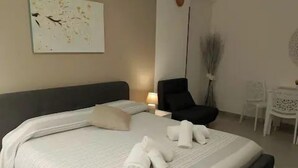 Uma cama bem arrumada com lençóis e toalhas brancas, uma cadeira moderna e uma pequena mesa em um quarto bem iluminado.