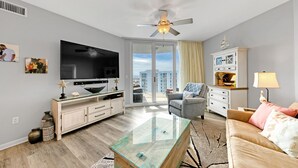 A modern living room with a flat-screen TV, a ceiling fan, a glass coffee table, a sofa, and a view of the city through a sliding glass door. - Palms of Destin|12th flr Jr Suite-Lagoon Pool|Views (Destin)
