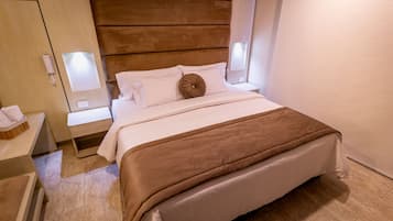 Una habitación de hotel con cabecera de madera, cama con ropa blanca, manta marrón y lámparas de noche.