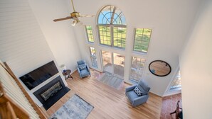 A spacious living area with a fireplace, a ceiling fan, and large windows offering a view of the outdoors. - Riverfront Retreat 7mil from MD Renaissance Fest! (Riva)