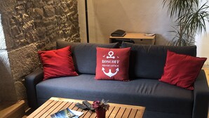 A cozy living room with a stone wall, a dark blue sofa, red cushions, a wooden coffee table with magazines, and a plant.
