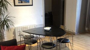A dining area with a round table, six chairs, a red cushion on a black sofa, a framed painting on the wall, and a potted plant.