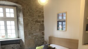 A bedroom with a stone wall, a bed with patterned bedding, a lamp, and a window with a view of buildings.