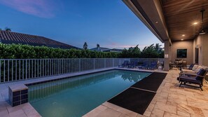 A swimming pool with a tiled deck, lounge chairs, and a covered seating area.