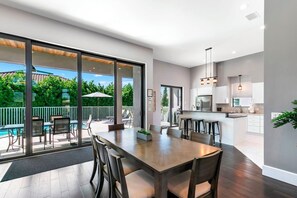 A modern kitchen with a central island, a dining area with a large table and chairs, and a view of a swimming pool and greenery outside.