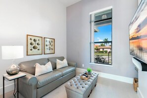 A modern living room with a grey leather sofa, a small side table with a lamp, a grey ottoman, and framed botanical prints on the wall. - Live in Luxury; Walk to the beach, shops and more (Marco Island)
