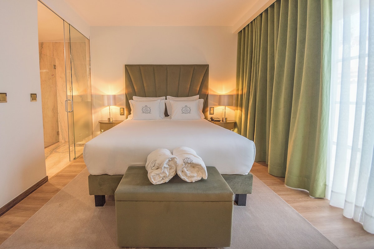Une chambre d’hôtel avec un grand lit, deux tables de chevet, un pouf vert et une cabine de douche en verre.
