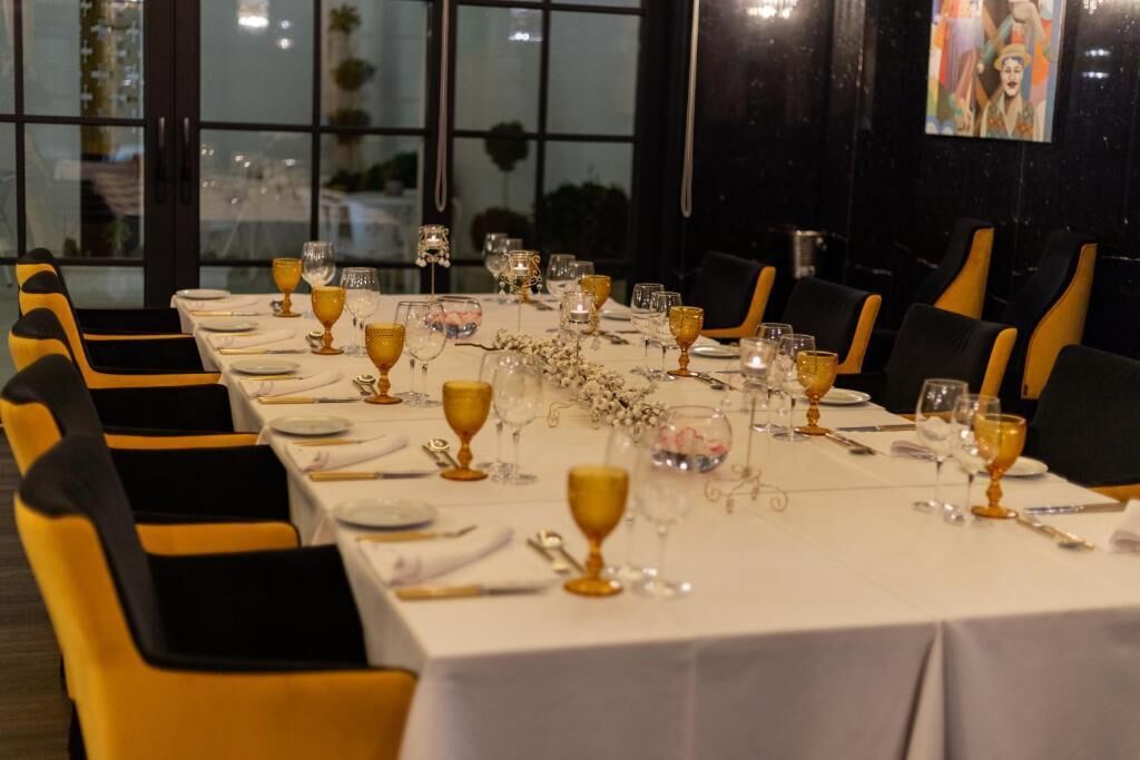 A long dining table set with gold-rimmed glasses, white tablecloth, and black and yellow chairs.