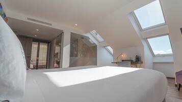 Un dormitorio moderno con una cama grande, tragaluces, un baño con puerta de vidrio y una pequeña kitchenette.