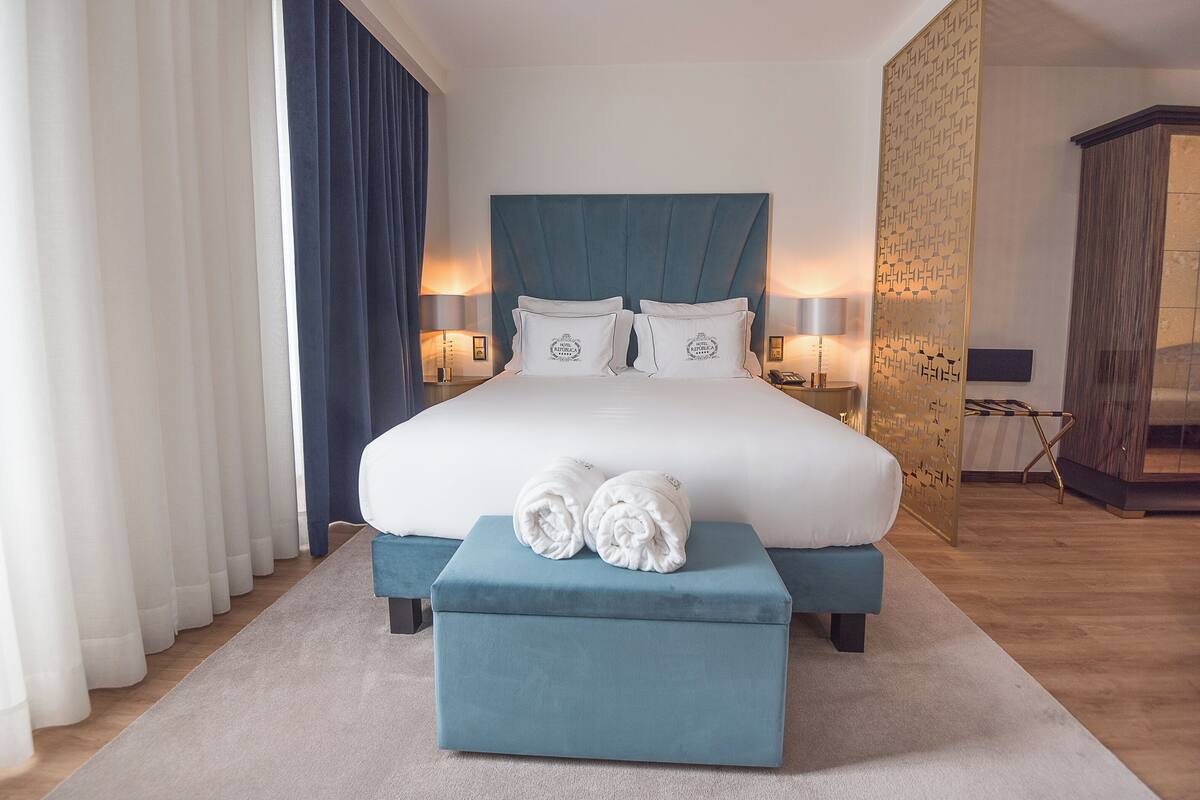 Une chambre d’hôtel moderne avec un grand lit, deux tables de chevet avec des lampes, une tête de lit bleue et un petit pouf avec des serviettes.