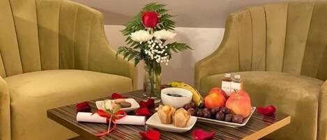 A dining table with a vase of flowers, fruits, and pastries in a room with two green upholstered chairs.