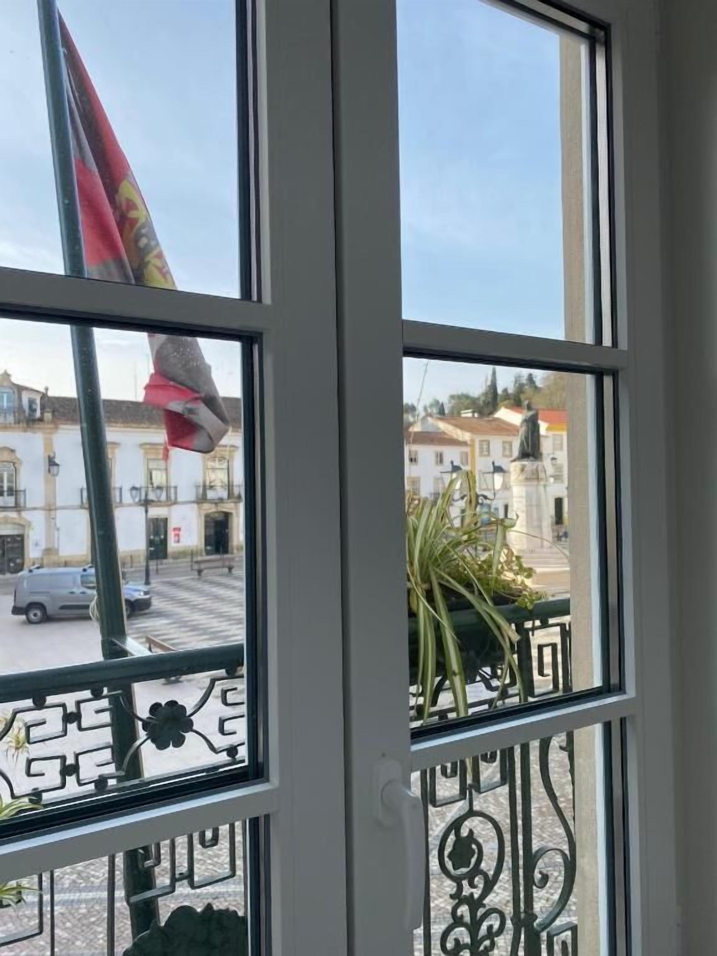 Una ventana con vista a una calle, una bandera y la barandilla de un balcón.
