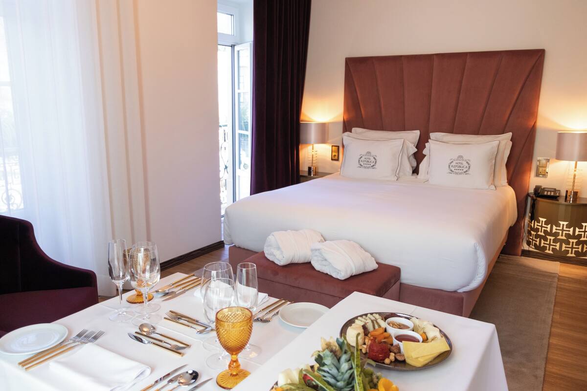Une chambre d’hôtel avec un grand lit, une table à manger dressée pour deux personnes et un plateau de petit-déjeuner contenant divers aliments.