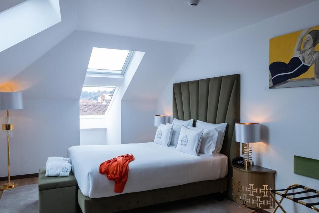A modern bedroom with a large bed, a skylight, a painting, and a bedside table with a lamp.