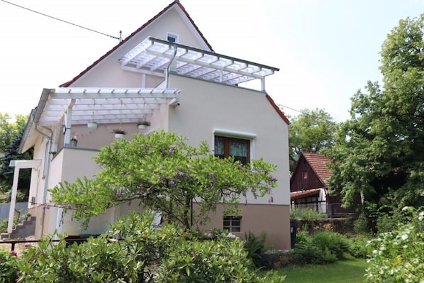 Une maison à deux étages avec un toit en pente, une pergola dotée d’un toit en verre et un jardin où poussent différentes plantes et arbres.