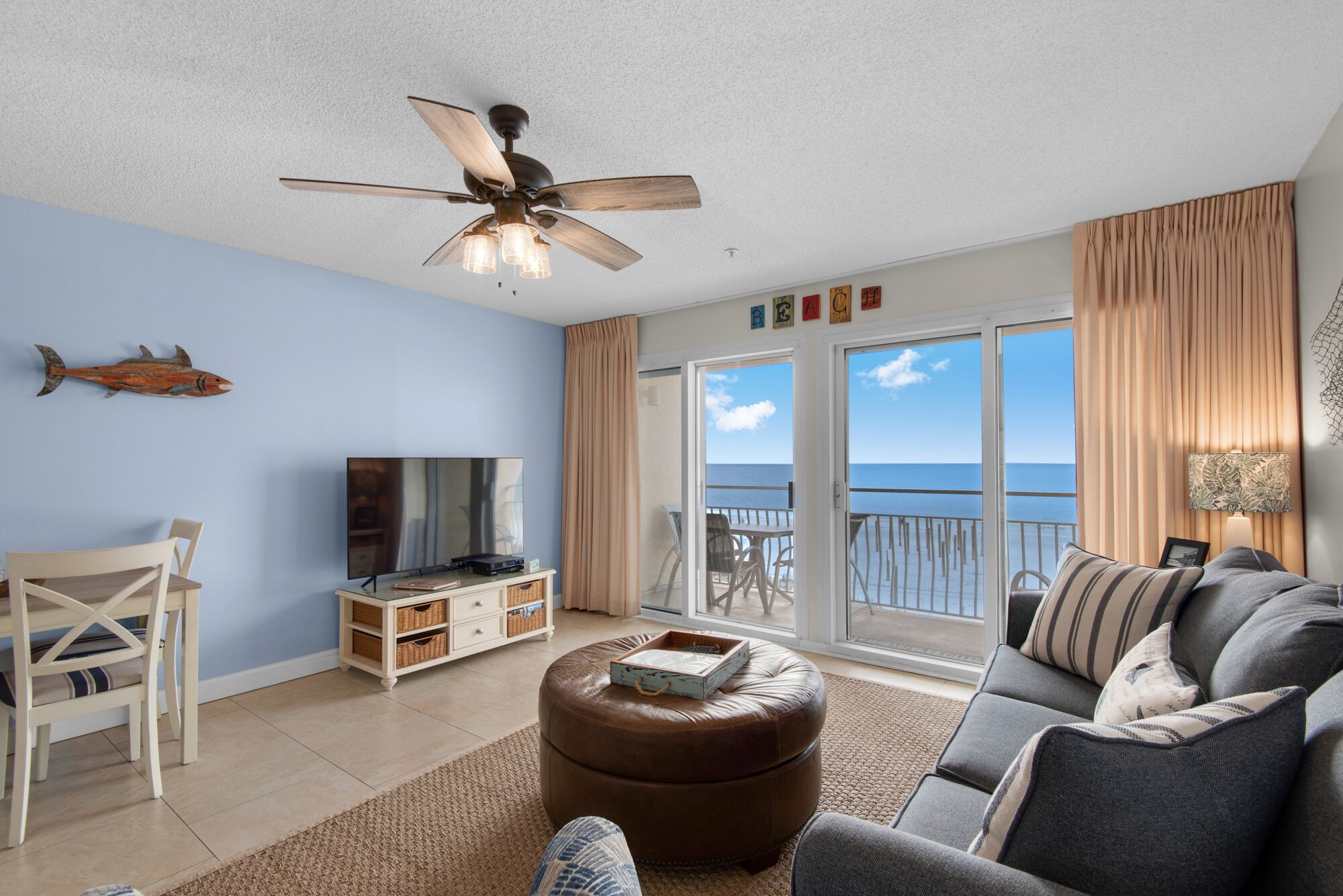 A living room with a TV, a ceiling fan, a sofa, and a view of the ocean through large sliding doors.