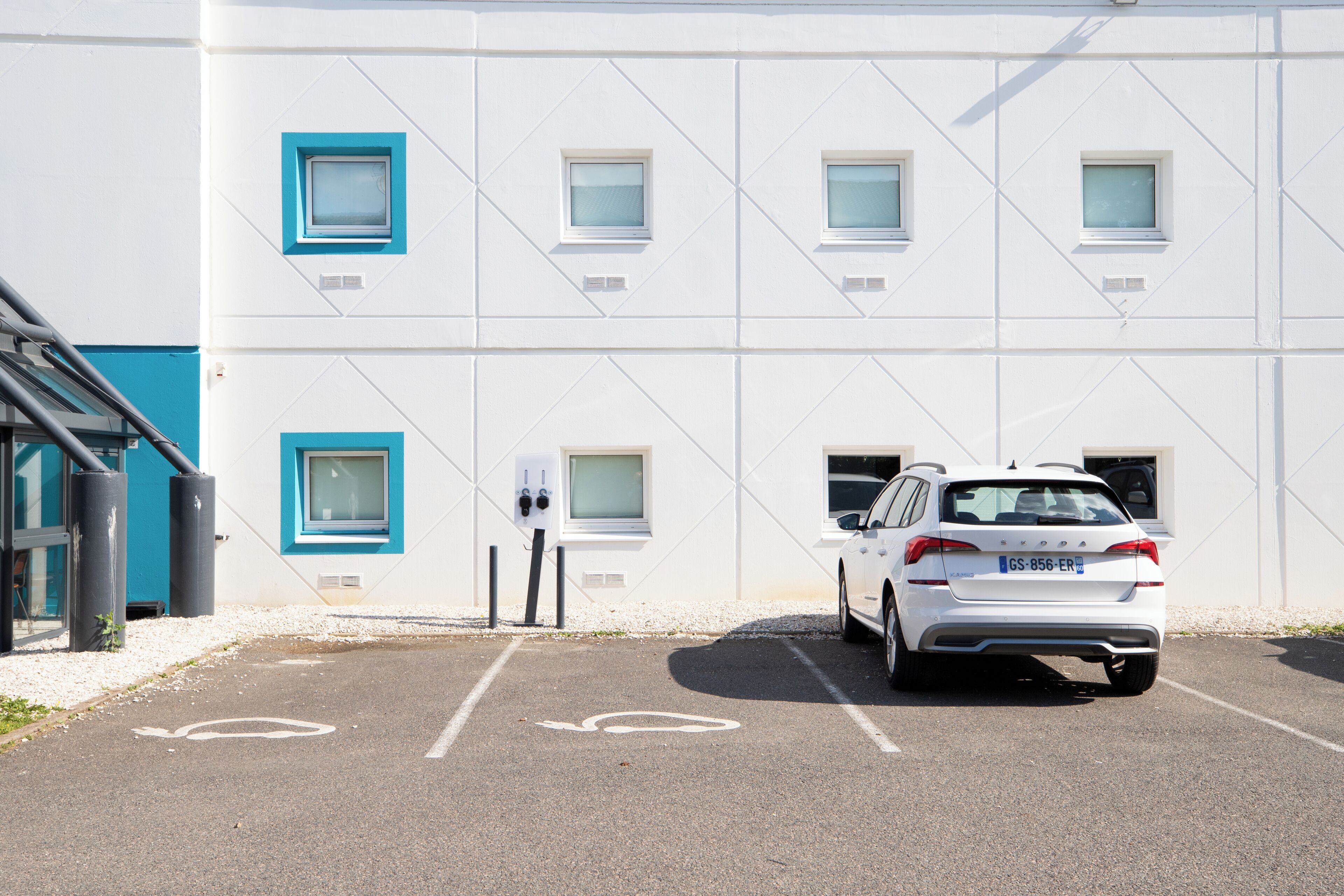 Un bâtiment blanc doté de nombreuses fenêtres et d’un espace de stationnement où se trouve une voiture blanche.