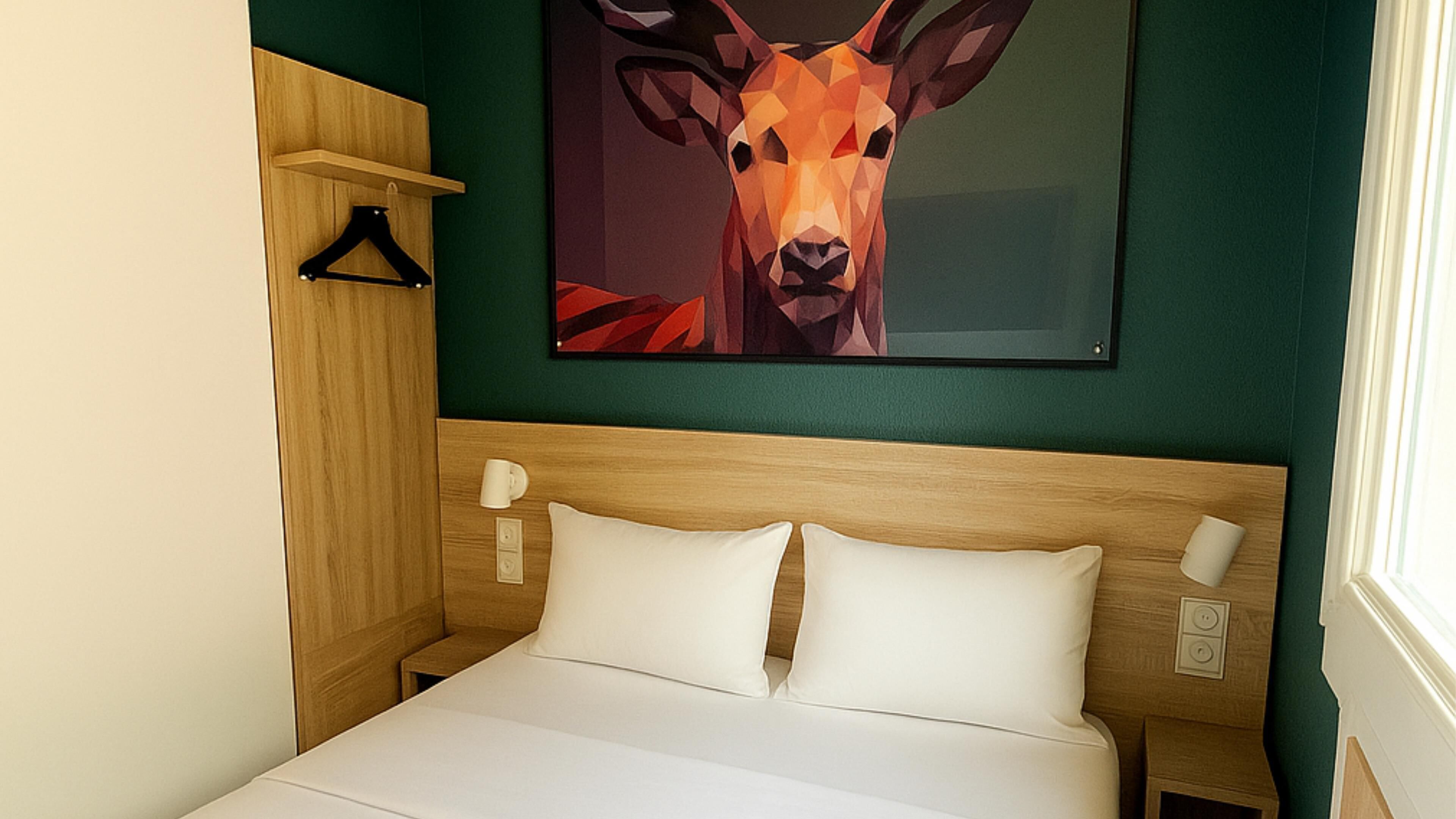A hotel room with a bed, a wooden headboard, a wall-mounted painting of a deer, a bedside table with a lamp, and a window with curtains.