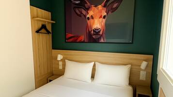 Une chambre d’hôtel avec un lit, une tête de lit en bois, un tableau représentant un cerf accroché au mur, une table de chevet avec une lampe et une fenêtre avec des rideaux.