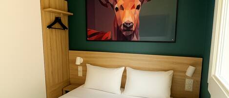 A hotel room with a bed, a wooden headboard, a wall-mounted painting of a deer, a bedside table with a lamp, and a window with curtains.