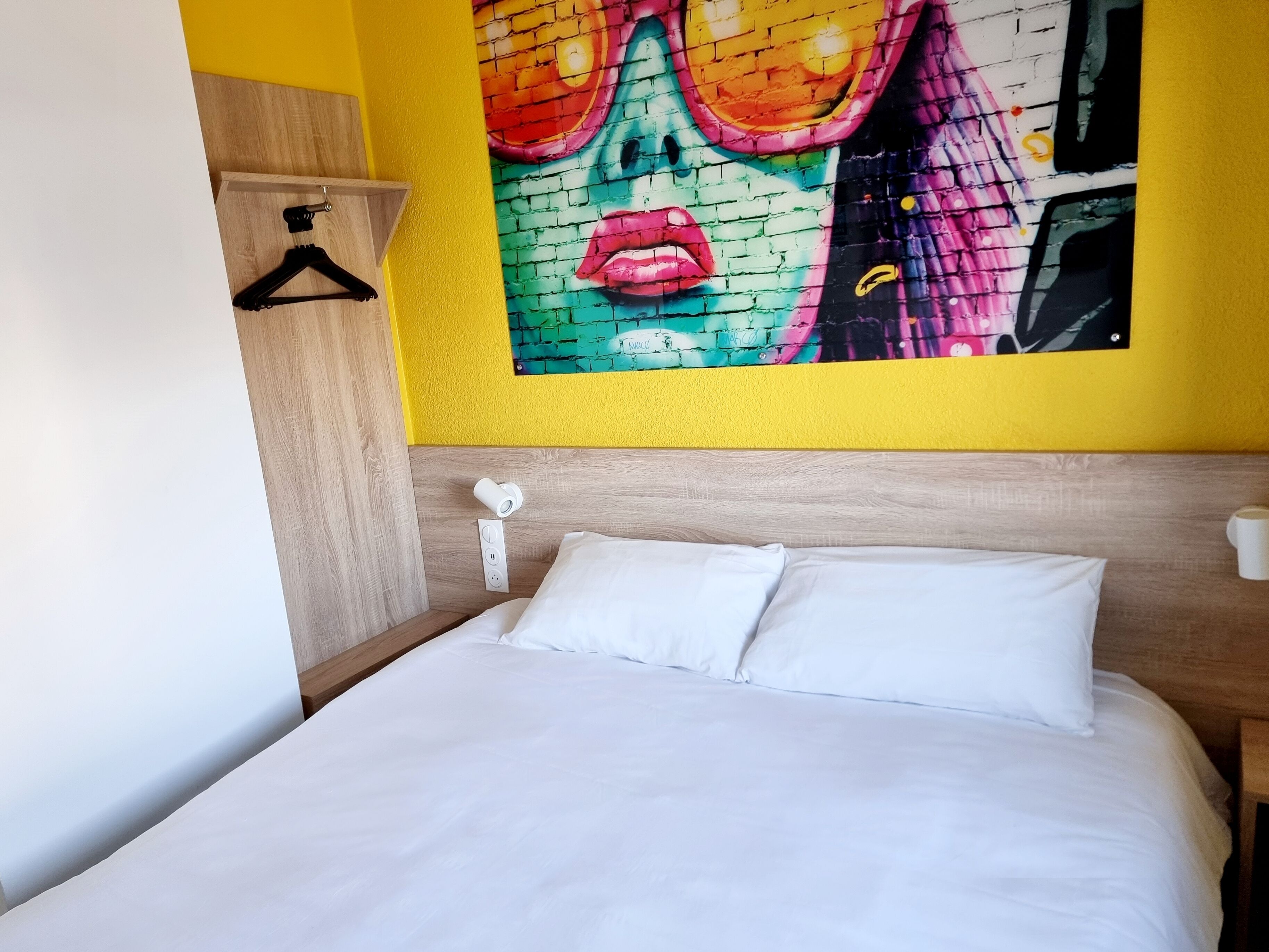 A hotel room with a bed, a wall-mounted light, a power outlet, and a colorful mural on the wall.