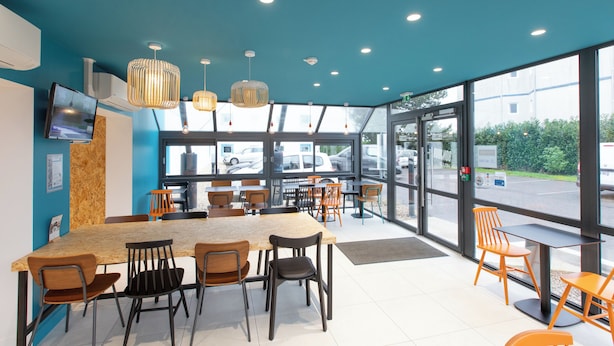 A modern restaurant with a large wooden table and chairs, a TV mounted on the wall, and large windows offering a view of the parking lot. - Kyriad ECO - Nevers Nord Varennes Vauzelles (Varennes-Vauzelles)