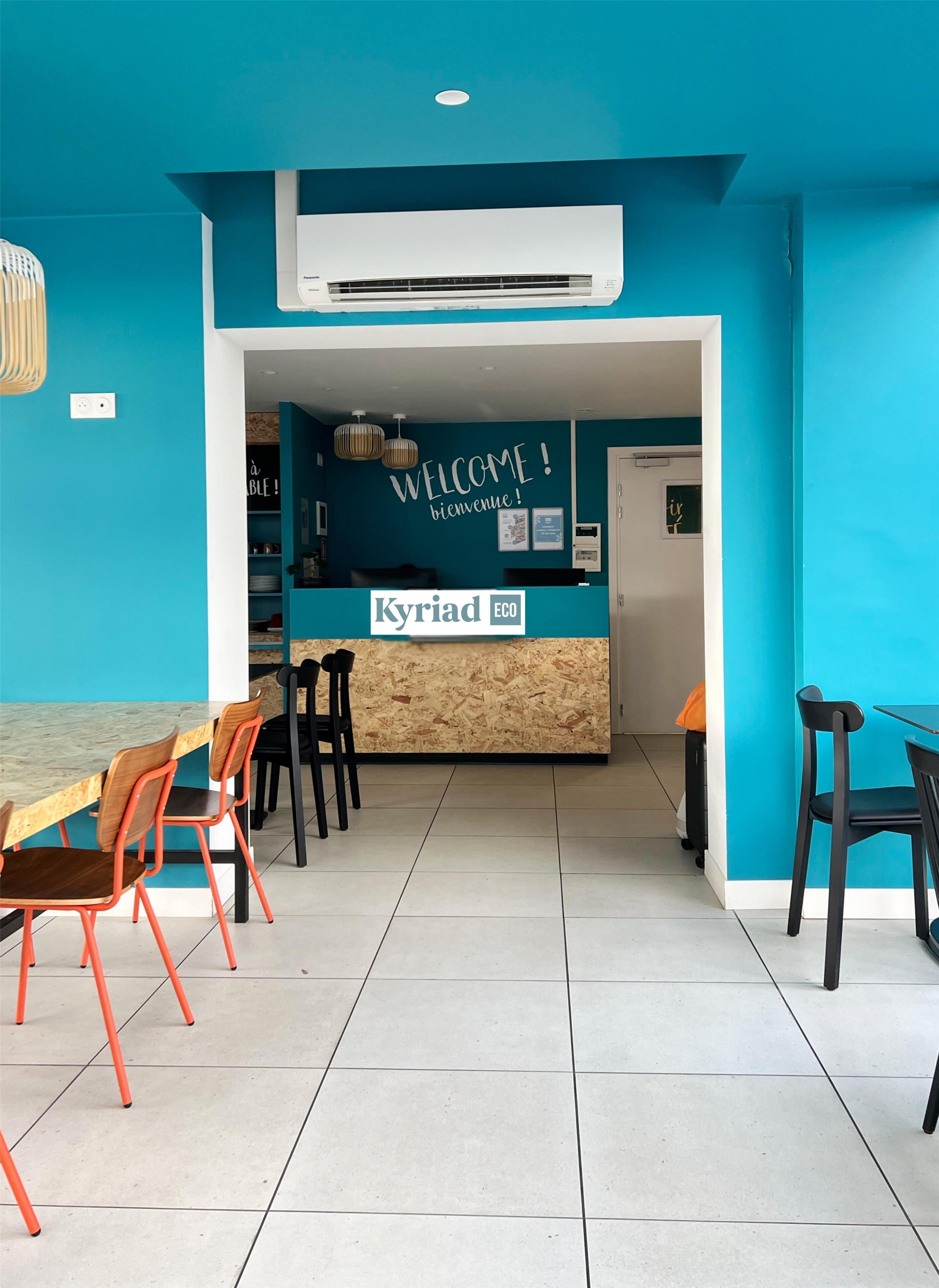 A reception area with a blue wall, a wooden reception desk, and a sign that reads "Kyriad ECO". There are chairs and a table in the foreground.