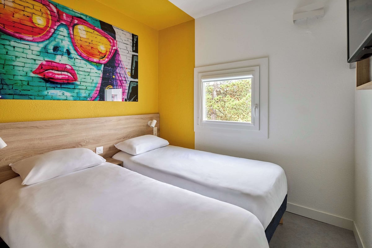 A hotel room with two beds, a colorful mural on the wall, a window with a view of trees, and a TV mounted on the wall.