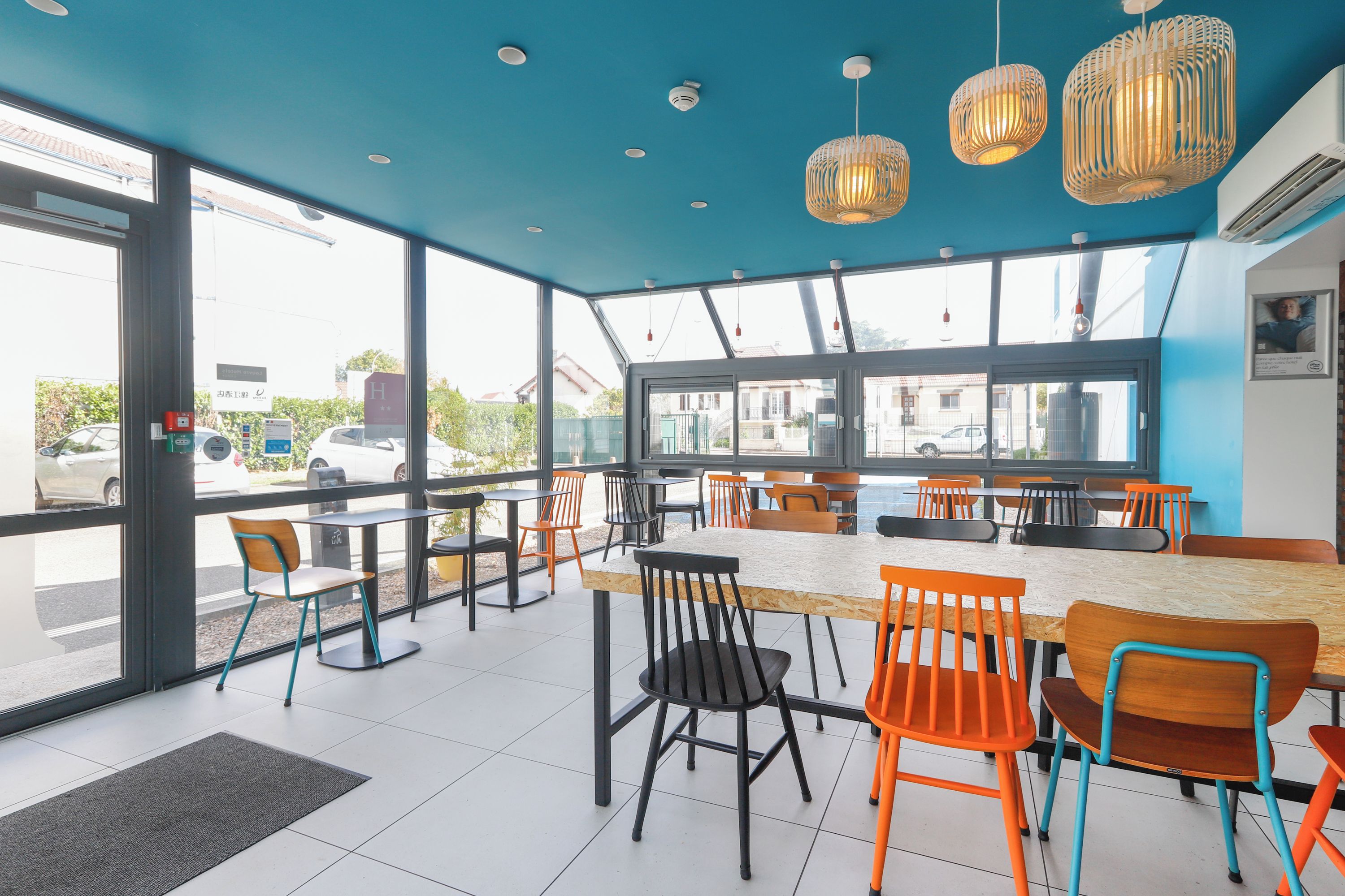 A modern cafe with large windows, outdoor seating, and a blue ceiling.