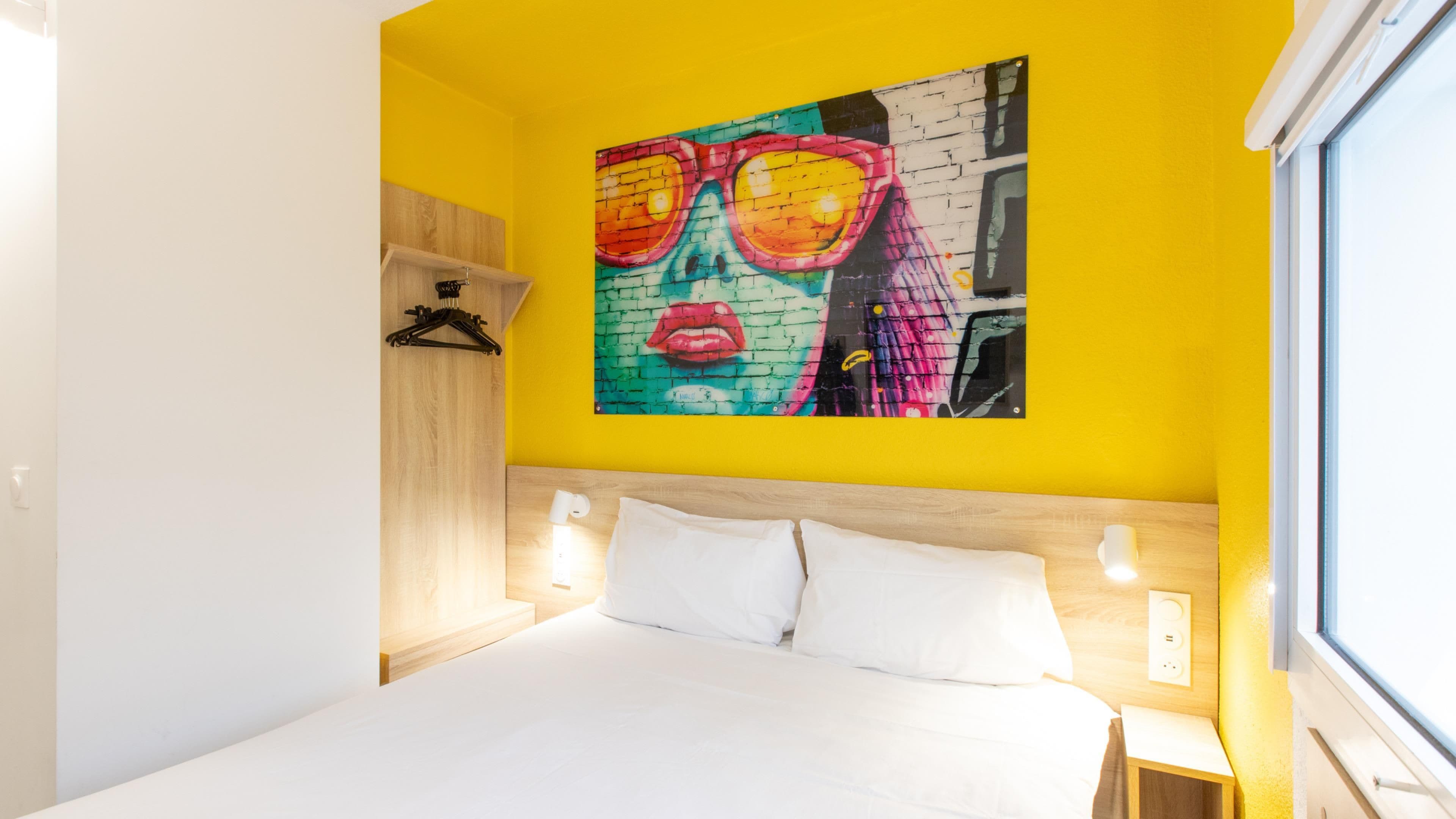 Ein Hotelzimmer mit einem Bett, einem Wandbild, einem hölzernen Kopfteil, Nachttischlampen und einem Fenster.