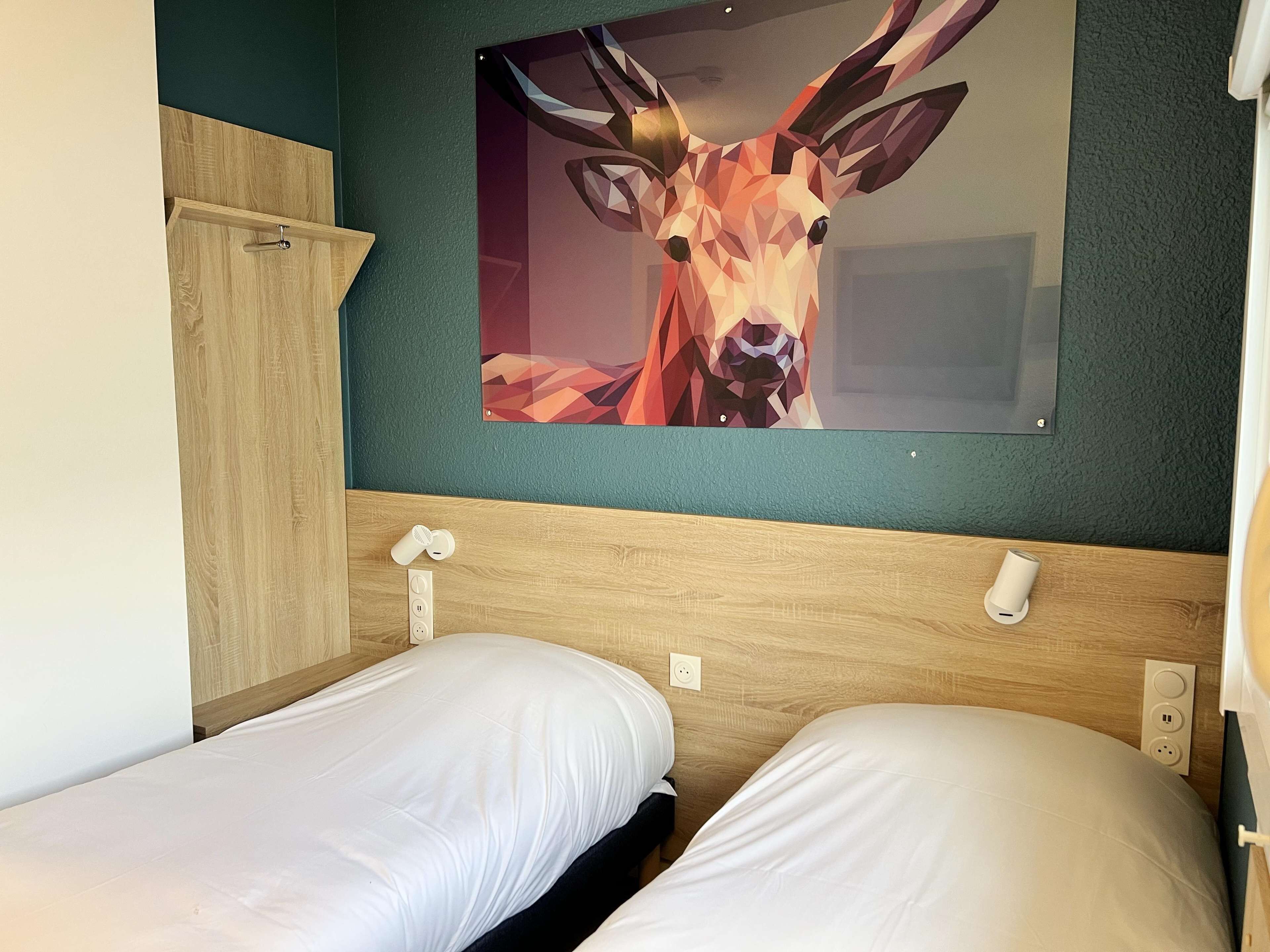 A hotel room with two single beds, a wooden headboard, a wall-mounted painting of a deer, and a small shelf with a cup and a container.