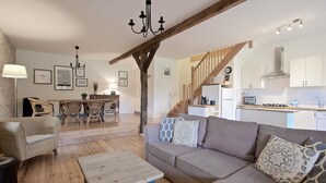 A spacious living area with a wooden floor, a sofa, a coffee table, and a dining area with chairs and a table. A staircase leads to an upper level with a kitchen.