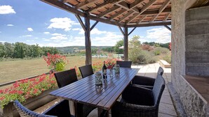 A covered patio with a wooden table and wicker chairs, overlooking a scenic landscape.