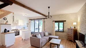 A spacious living area with a sofa, a coffee table, and a TV. There is a kitchen in the background with white cabinets and a stove.