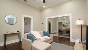 Un salon moderne avec un canapé d’angle beige, un miroir rond, une table basse en bois et un coin repas comprenant une table et des chaises en bois.