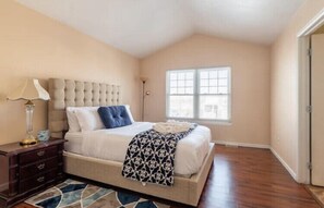 A bedroom with a tufted headboard, a bed with white linens and a patterned throw, a nightstand with a lamp, and a large window.
