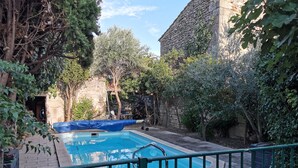Une piscine entourée de murs en pierre et de verdure, avec un espace abrité et une rambarde en métal.