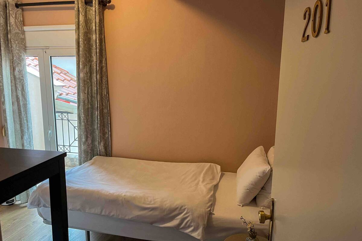 A single bed with white bedding, a small bedside table, a vase with flowers, and a door with the number 201.