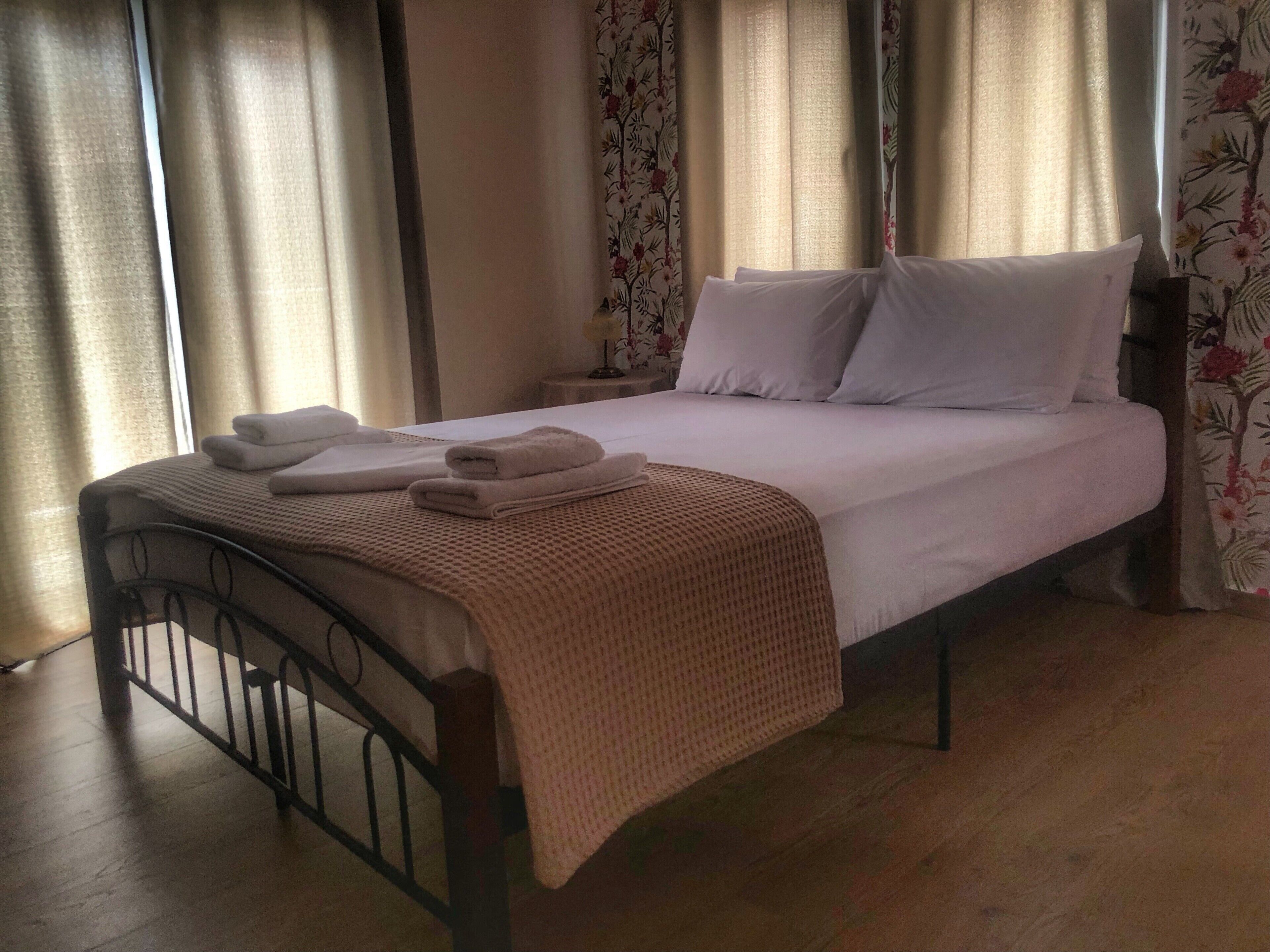A neatly made bed with white linens and a beige blanket, a wooden headboard, and floral curtains.