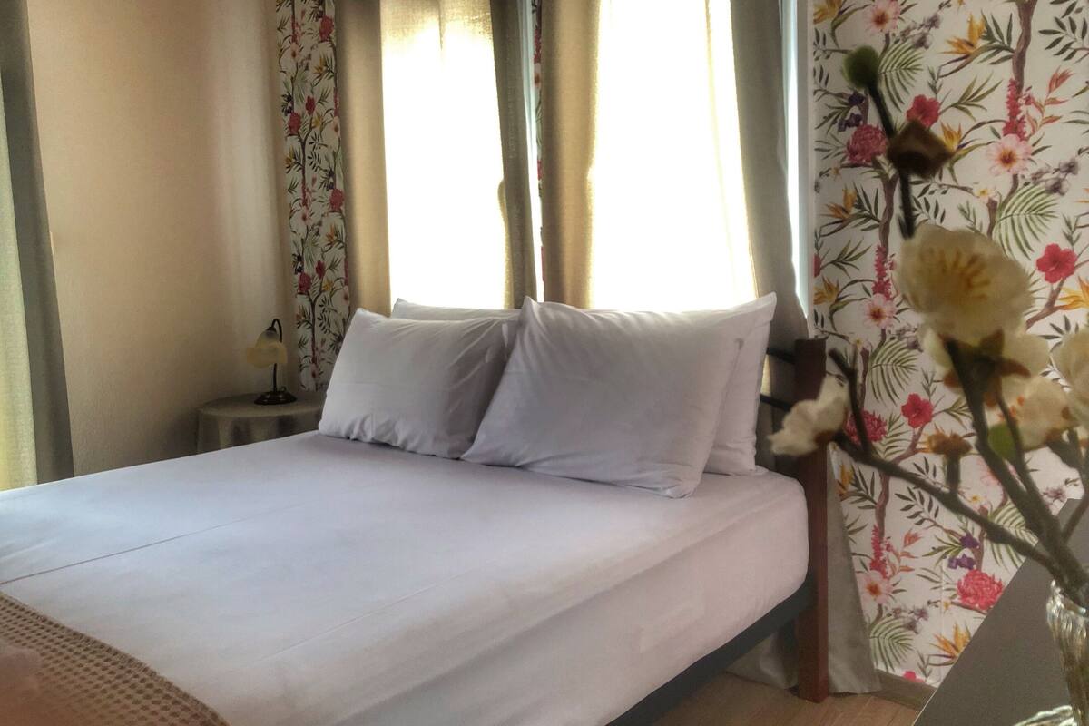A bed with white linens in a room with floral wallpaper and a window with curtains.