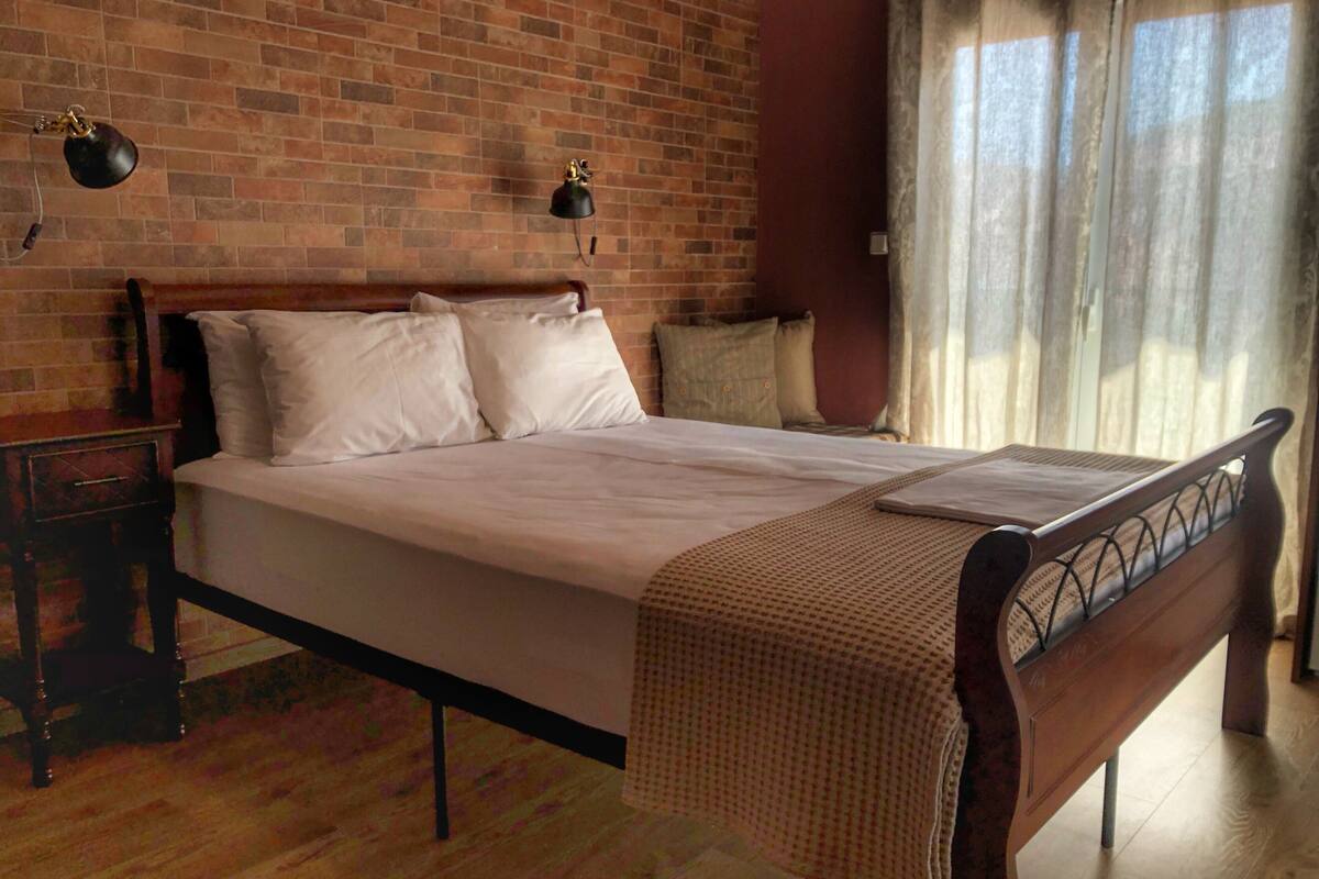 A neatly made bed with a wooden headboard, a bedside table with a lamp, and a brick wall.