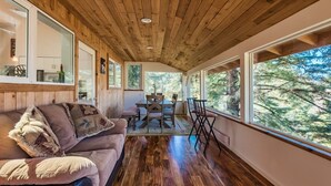 Ein gemütliches Blockhaus-Interieur mit Holzbalkendecke, einem Essbereich mit Tisch und Stühlen sowie großen Fenstern mit Blick auf den Wald.
