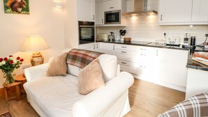 A modern kitchen with white cabinets, a microwave, and a coffee machine. There is a white sofa with a plaid throw blanket and a small table with a lamp and a vase of flowers. - The Coach House (Bishop Auckland)
