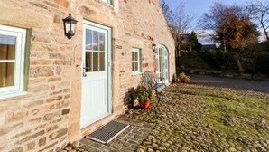 A stone building with a white door, a window with a green frame, and a small garden area with a bench and potted plants. - The Coach House (Bishop Auckland)