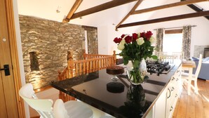A kitchen with a stone wall, wooden beams, a black countertop, white chairs, and a vase with flowers. - The Barn (Bishop Auckland)