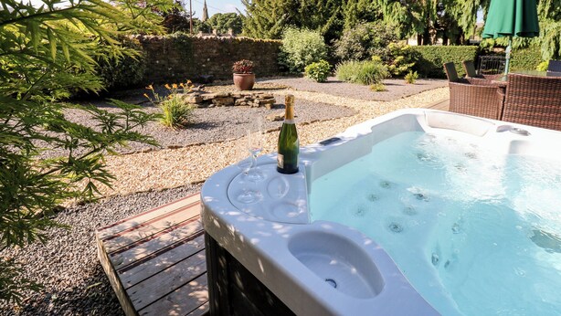 A hot tub with a bottle of champagne, surrounded by a garden with various plants and a stone wall. - The Barn (Bishop Auckland)