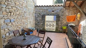 Une terrasse dallée de pierres, avec une table ronde en bois et des chaises, un portail en métal et une petite chaise verte.