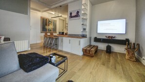 A modern living room with a sofa, coffee table, and a TV mounted on the wall. There is a kitchen area with a dining table and chairs.