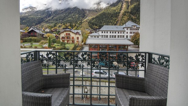 A balcony with wicker furniture overlooking a town with buildings and mountains in the background.